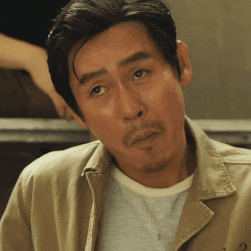 a man with a beard wears a tan jacket and a white shirt