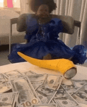 a doll in a blue dress is sitting on a table surrounded by 100 dollar bills .