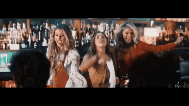 three women are dancing in a bar in front of a bar full of bottles of alcohol .