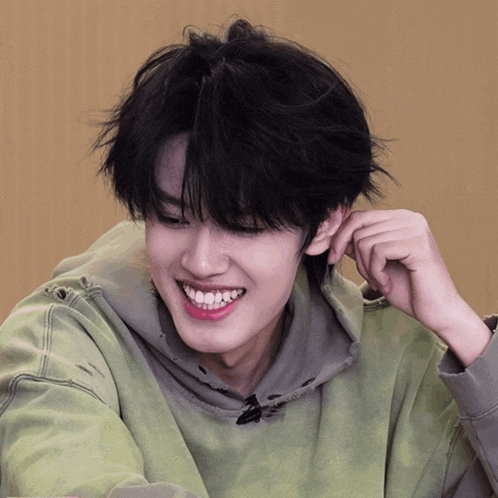 a young man in a green hoodie smiles with his hand on his ear