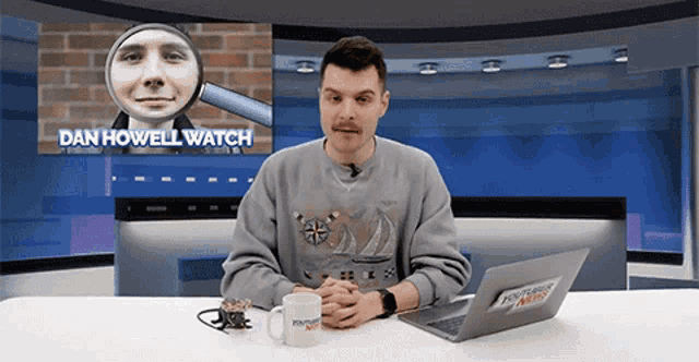 a man sitting at a desk with a laptop and a mug with the name dan howell watch behind him
