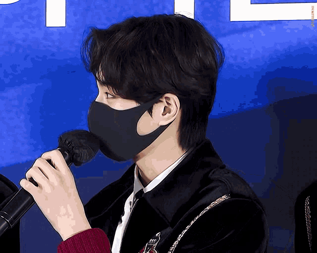 a man wearing a black mask holds a microphone in front of a blue background