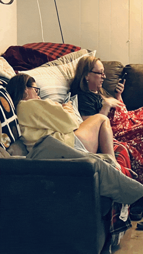 two women are sitting on a couch looking at their cell phones
