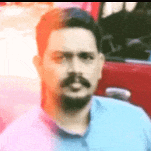 a man with a beard and mustache is wearing a blue shirt and standing in front of a red car .