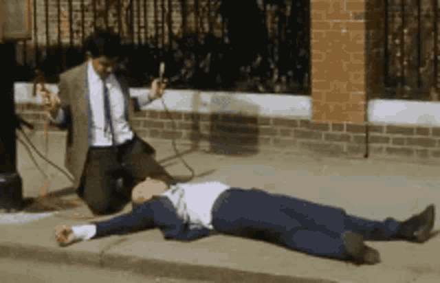 a man in a suit and tie is kneeling down next to a man laying on the sidewalk
