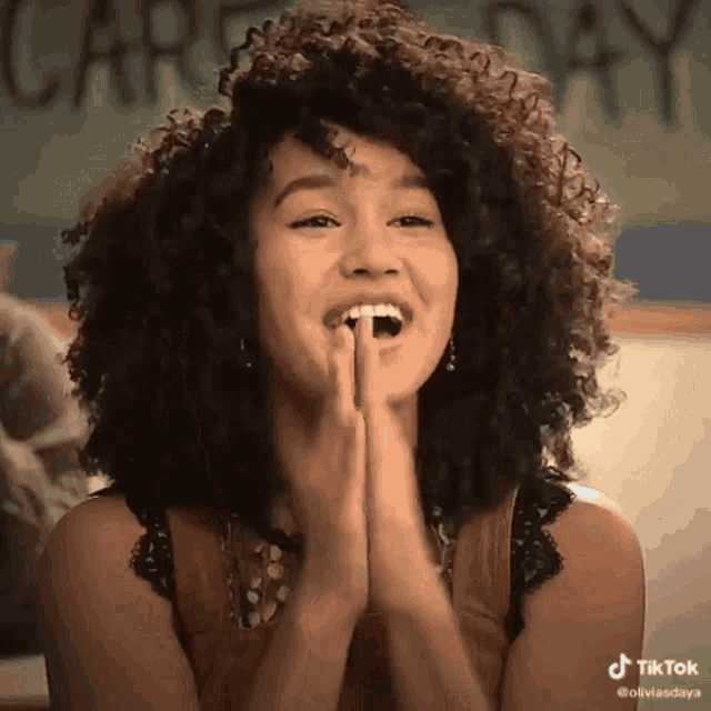 a woman with curly hair is smiling with her hands folded in front of her face