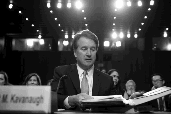 a man in a suit and tie sits at a table with a sign that says m. kanaugh
