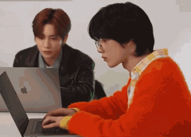 a man in an orange sweater is typing on a laptop next to another man in a black jacket