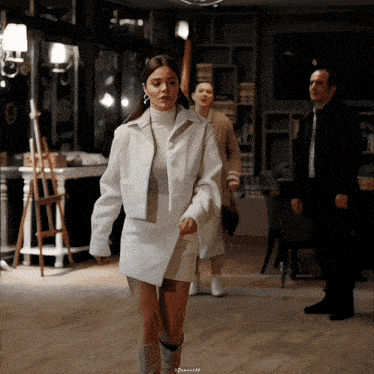 a woman in a white coat is walking in a room with a man in a suit behind her