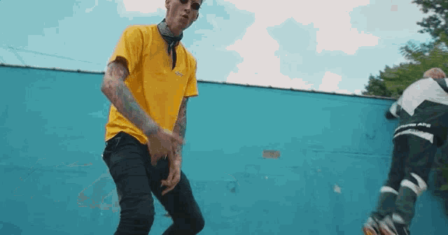 a man in a yellow shirt is standing in front of a blue wall while another man is rollerblading .