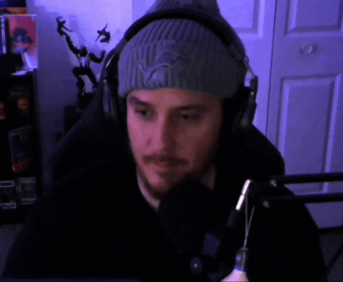a man wearing a beanie and headphones looks at the camera in front of a microphone