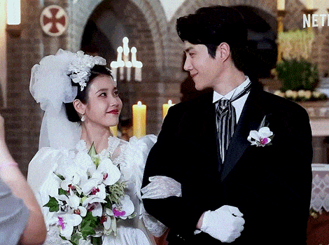 a bride and groom standing next to each other in front of a church with a cross on the wall behind them