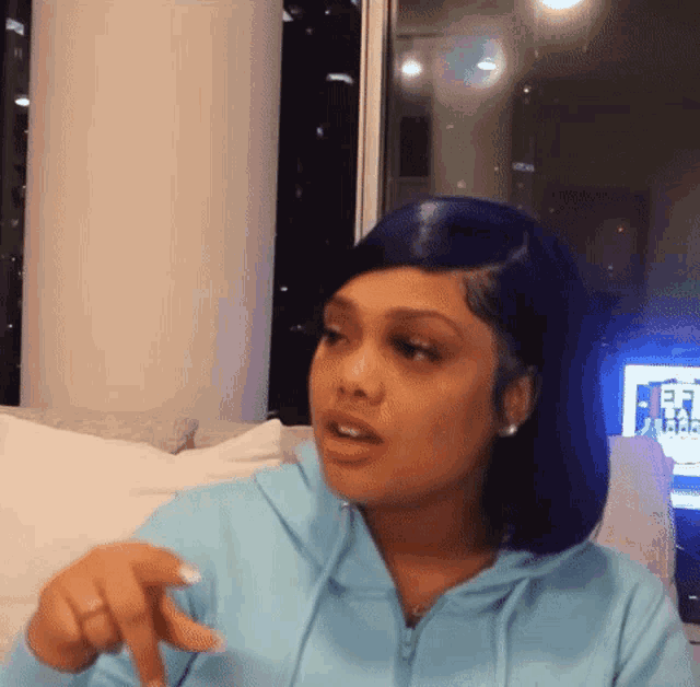 a woman in a blue hoodie is sitting on a couch and pointing at the camera .