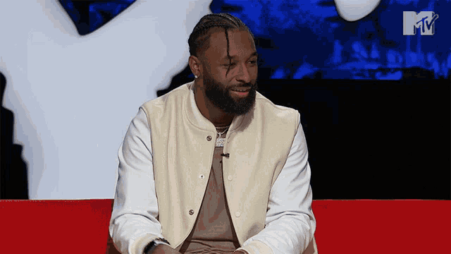 a man in a white jacket sits on a red couch with a mtv logo behind him