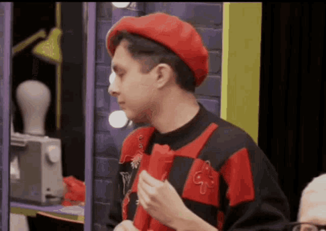 a man wearing a red beret and a red and black shirt is standing in front of a sewing machine .