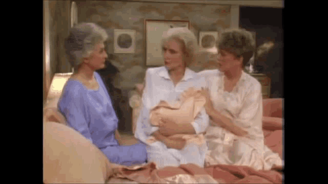 three older women are sitting on a bed talking to each other in a living room .
