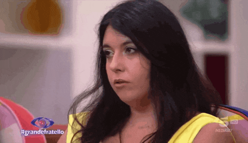 a woman in a yellow shirt is sitting in front of a sign that says #grandefratello