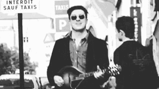 a black and white photo of a man playing a guitar in front of a sign that says interdit sauf taxis