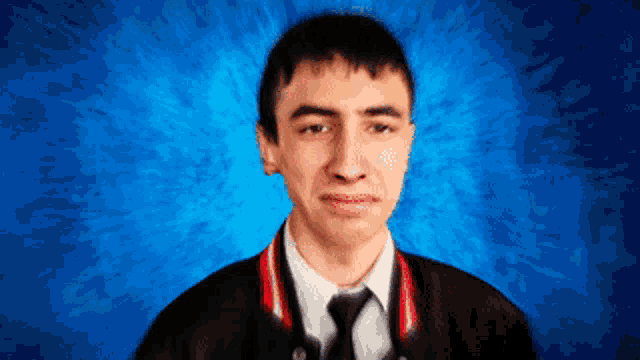 a young man wearing a black jacket and tie looks at the camera with a blue background