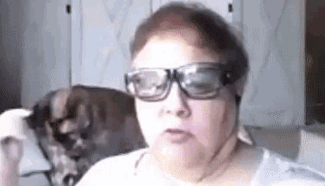 a woman wearing glasses is sitting in a chair with a dog in the background .