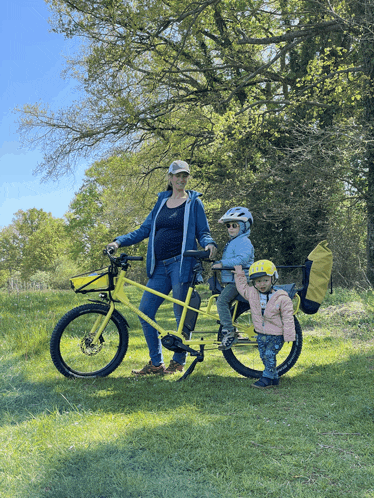 a woman is standing next to a yellow bicycle with two children on it