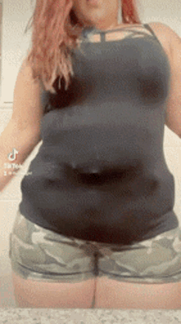 a woman in a black tank top and camo shorts is standing in front of a wall .