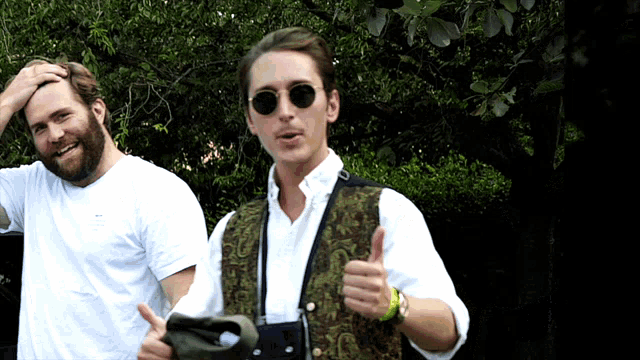 a man wearing sunglasses gives a thumbs up while standing next to another man