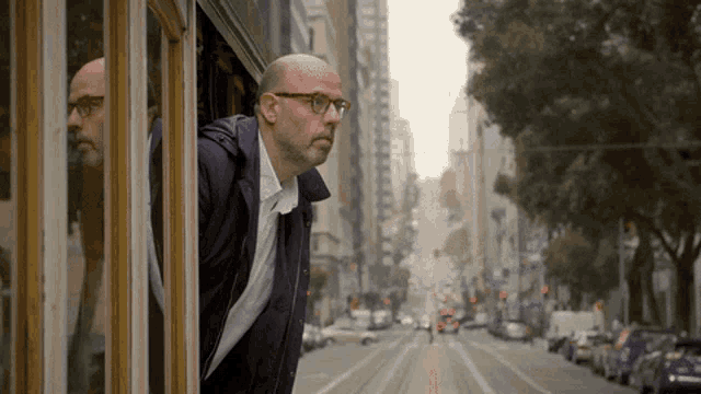 a man wearing glasses looks out a window at a city street