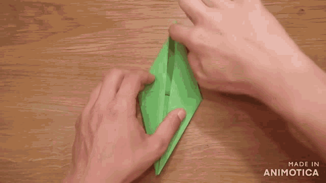 a person folding a piece of green paper on a wooden table with the words made in animatica on the bottom