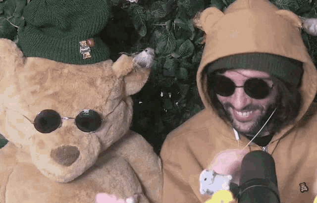a man wearing a hoodie and sunglasses is standing next to a teddy bear wearing a green hat