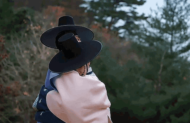 a man wearing a black hat and a pink blanket stands in front of a forest