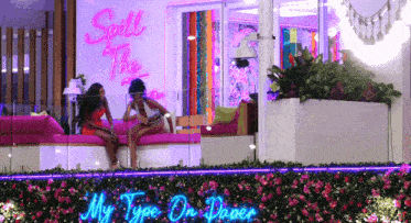 two women are sitting on a couch in front of a neon sign that says spill the tea