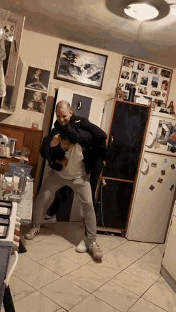 a man is carrying another man on his back in a kitchen with a picture of a tree on the wall