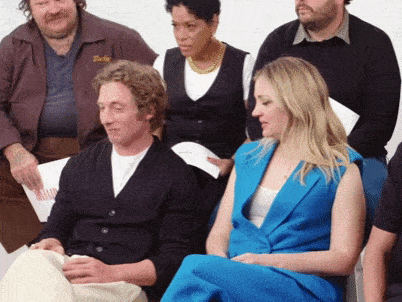 a woman in a blue dress sits in front of a group of people including a man wearing a jacket that says barley