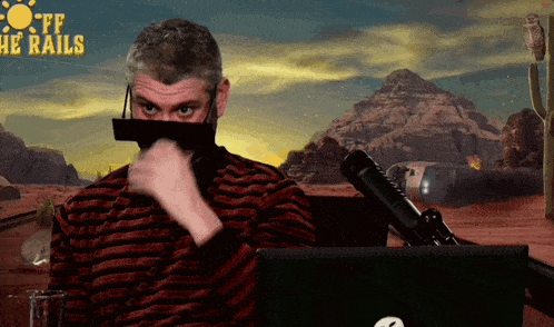 a man wearing a mask is sitting in front of a microphone with the words off the rails behind him