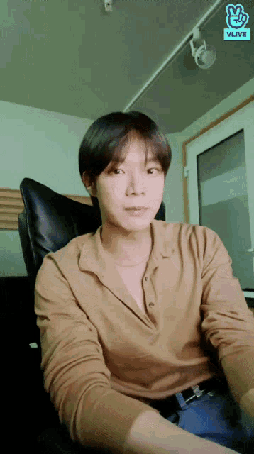 a man in a brown shirt is sitting in a chair with a vlive logo behind him