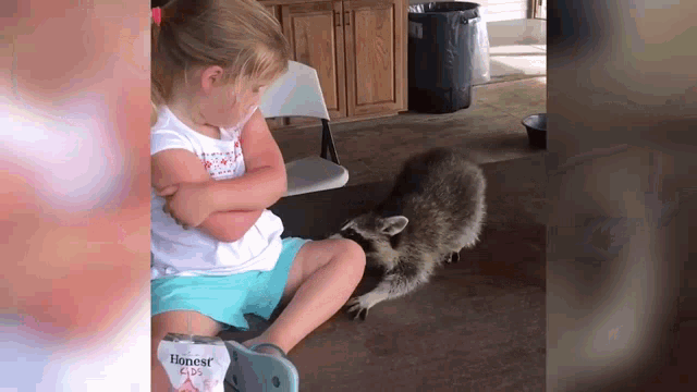 a little girl is sitting in a chair with her arms crossed while a raccoon looks on .