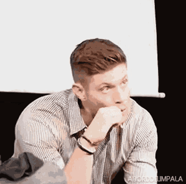 a man in a striped shirt is sitting in front of a white board with his hands on his chin .