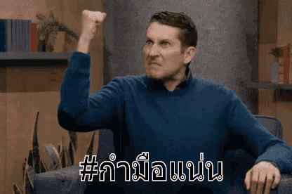 a man in a blue sweater is sitting on a couch with his fist in the air and a caption in another language .
