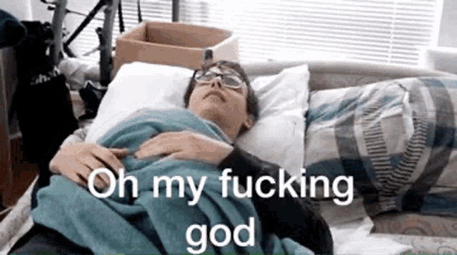 a man with glasses is laying in a bed with the words `` oh my fucking god '' written on it .
