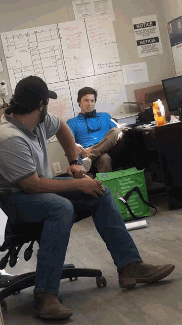 two men sit in front of a white board with a notice on it