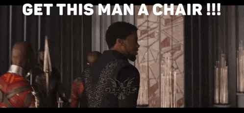 a man in a black jacket is standing in front of a wall with the words `` get this man a chair !! ''