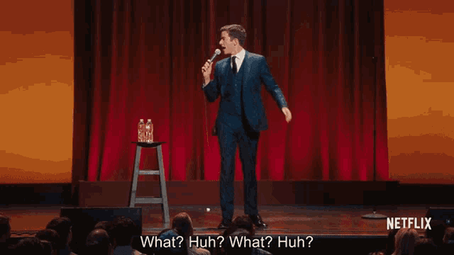 a man in a suit stands on a stage holding a microphone and says what huh