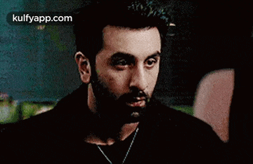 a man with a beard wearing a black shirt and a necklace is looking at a woman .