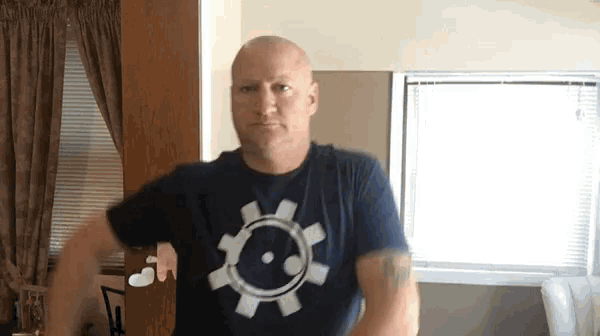 a bald man wearing a blue t-shirt with a gear on it is standing in front of a window .