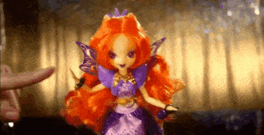a doll with orange hair and purple wings is being held by a person