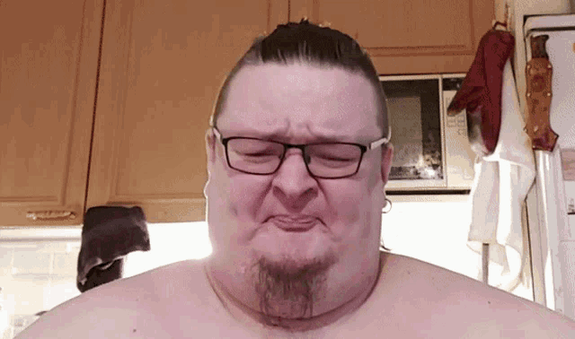 a shirtless man with glasses and a beard making a face in front of a microwave