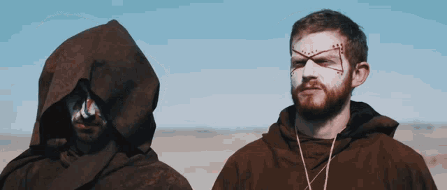 a man with a cross painted on his face stands next to another man with a hood