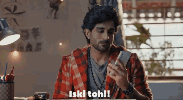 a man in a red plaid shirt is holding a cell phone and says iski toh