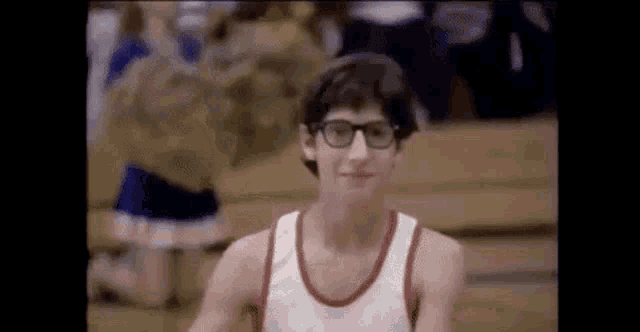a young man wearing glasses and a white tank top is sitting in a basketball court .
